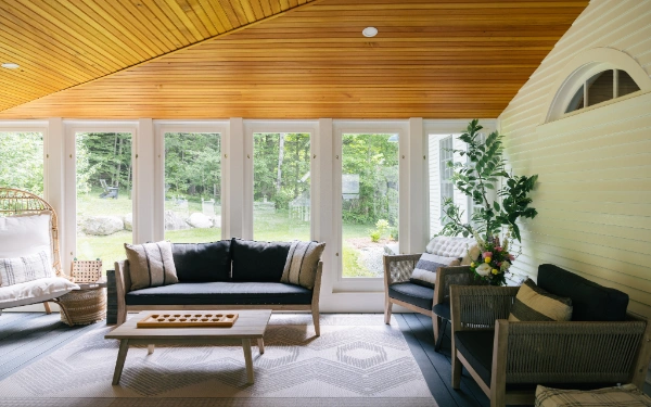 Sunroom seating area with woven lounge chairs, neutral cushions, wood-paneled ceiling, and floor-to-ceiling windows overlooking the yard